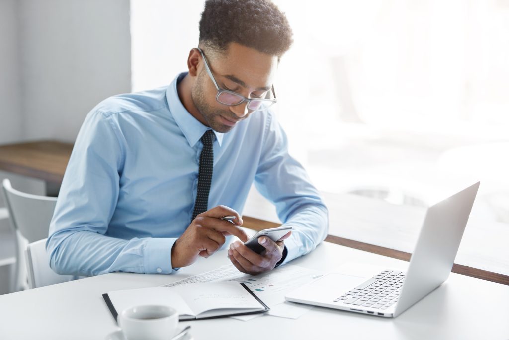 Picture of handsome African office worker having a few minutes break, texting sms on smart phone, sitting at his workplace with generic laptop computer, papers, notebook and mug of coffee on table